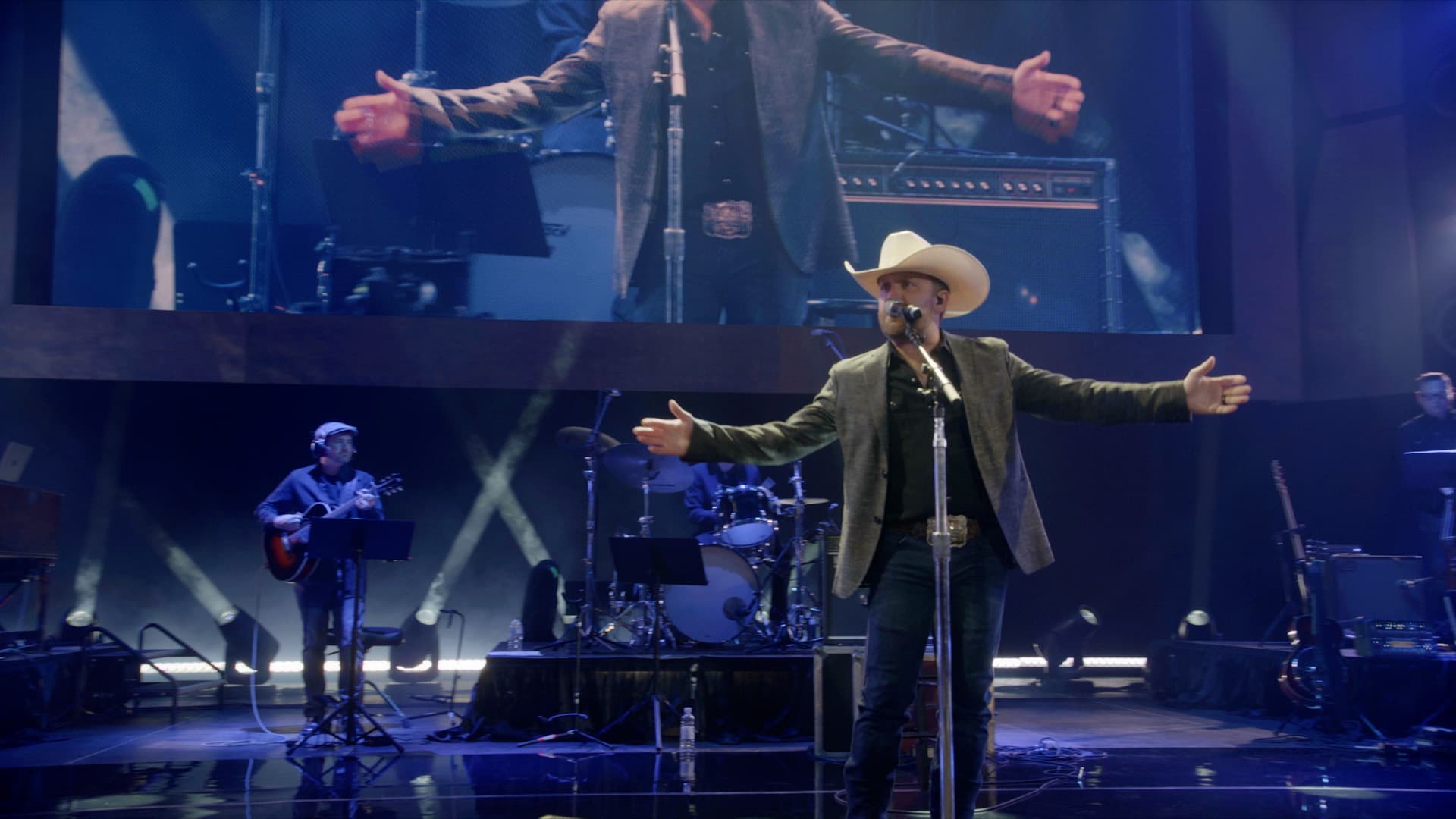 Justin Moore in a cowboy hat performs on stage with arms outstretched, while musicians play instruments behind him. A large screen displays his image in the background.