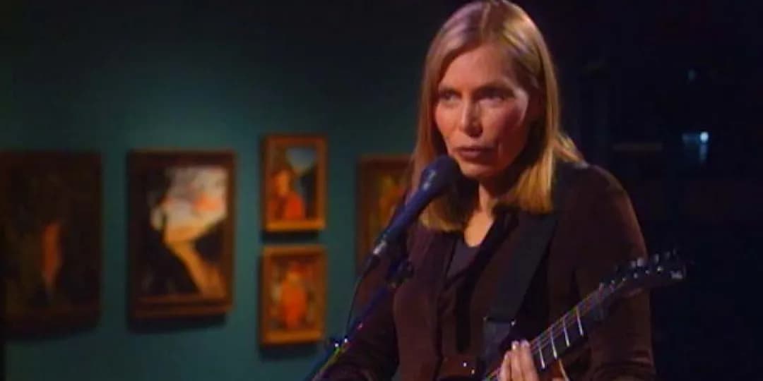 Joni Mitchell with long hair plays an electric guitar and sings into a microphone on a dimly lit stage, with framed artwork blurred in the background.