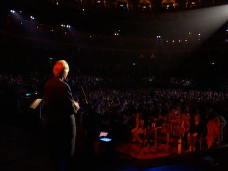 Sir George Martin stands on a stage under a spotlight, facing a large seated audience in a dimly lit auditorium.