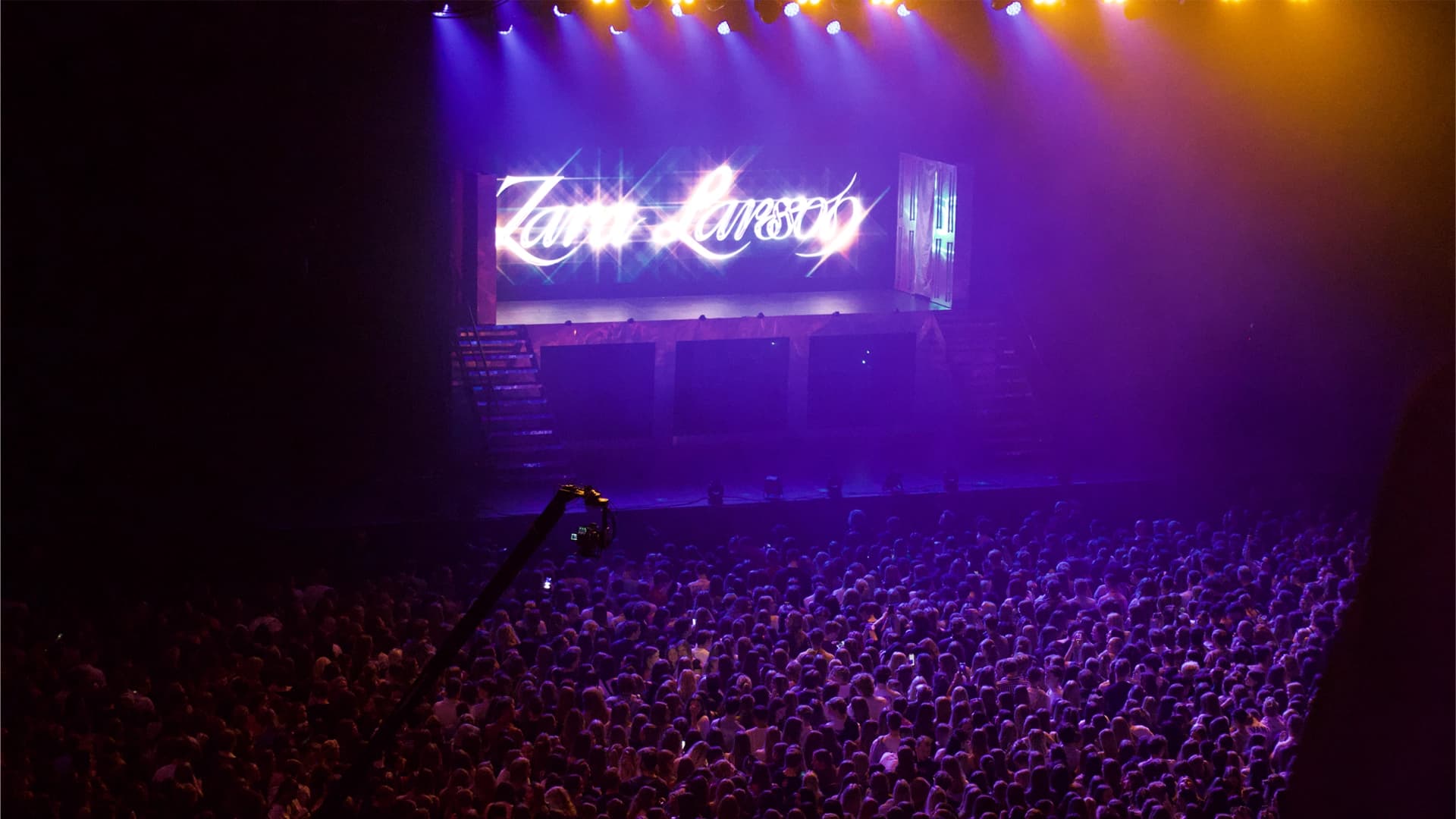 A large crowd of spectators at a concert venue with the stage in the background illuminated by a sign.
