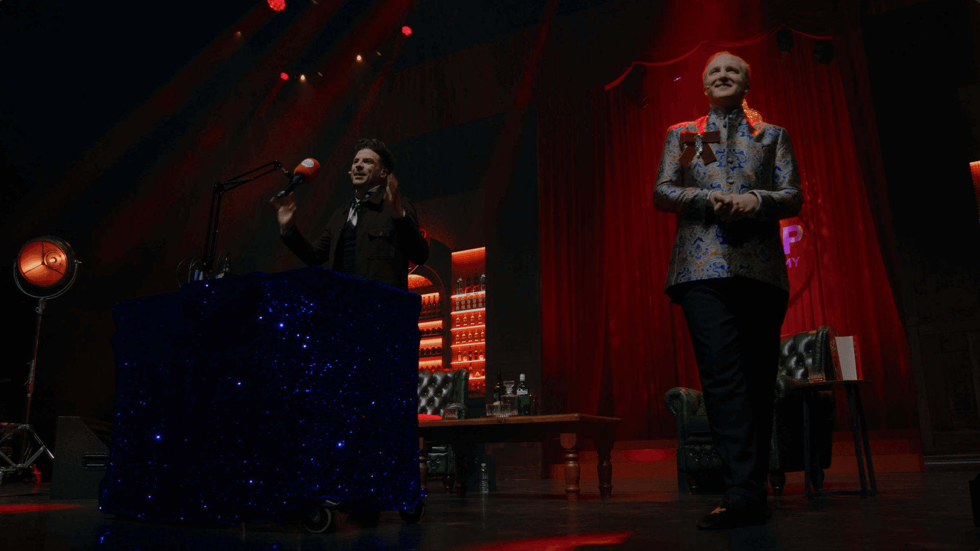 Two people stand on a stage with red curtains and dramatic lighting; one speaks at a sparkly podium, the other stands nearby, both facing the audience.