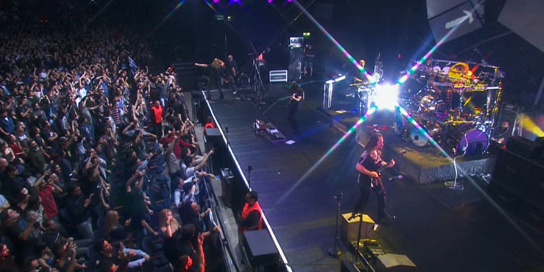Dream Theater performing on a stage with bright lights and a large crowd cheering in an indoor venue.