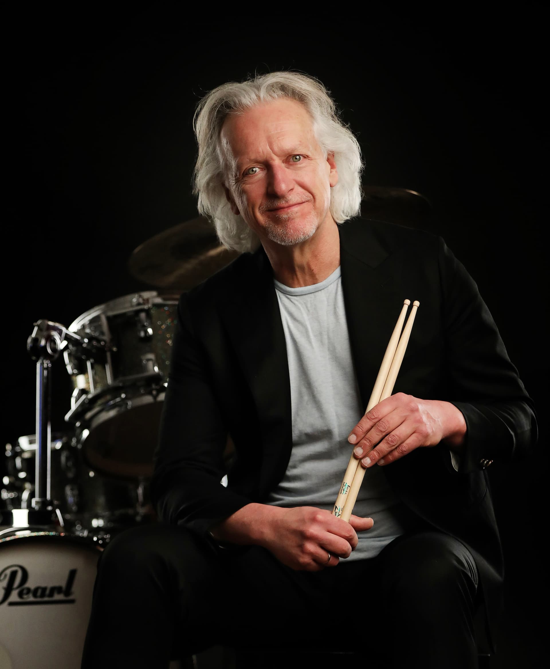 A man with white hair sits in front of a drum set, holding drumsticks and smiling at the camera. He is wearing a black jacket and a light gray shirt.