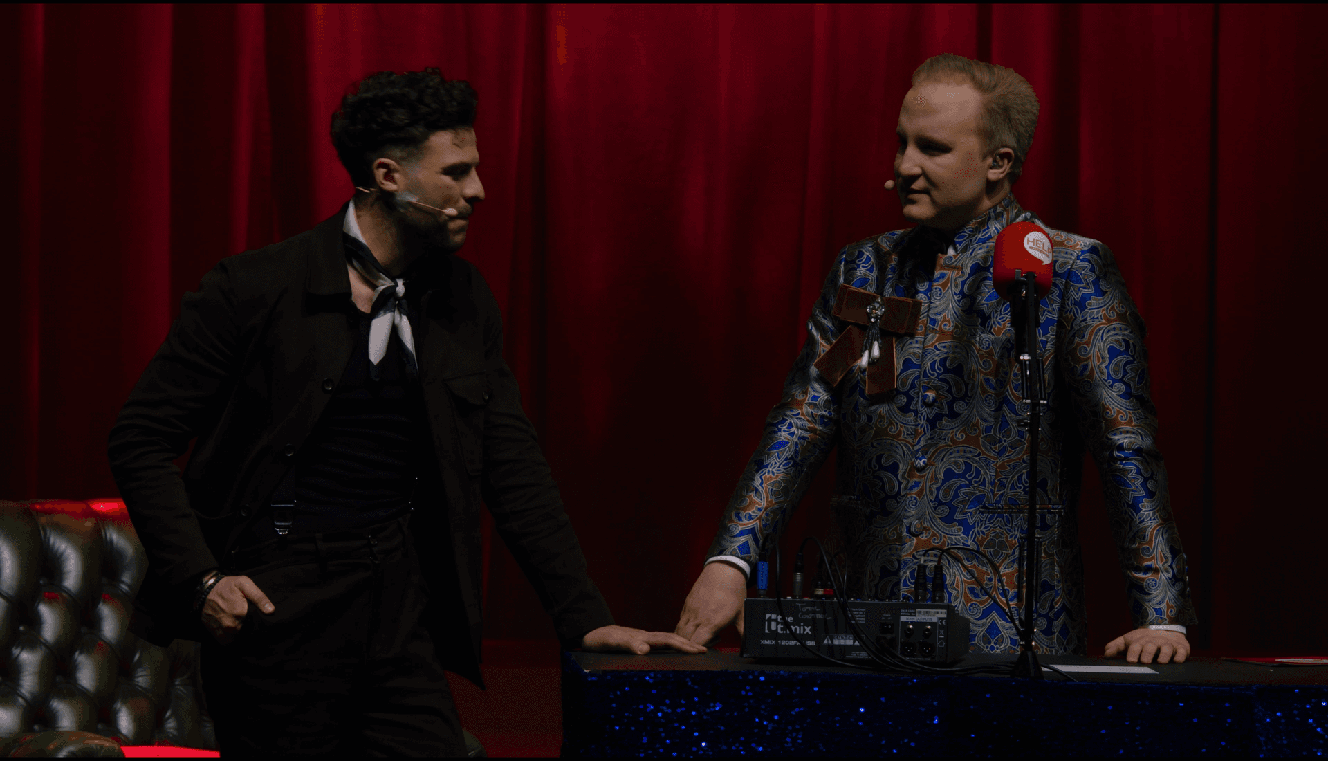 Two men on stage converse; one stands in dark attire with a scarf, the other in a patterned suit behind a table with microphones and audio equipment, red curtains in the background.