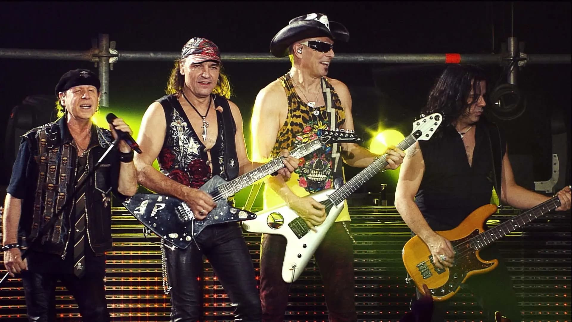 Scorpions perform energetically on stage, each playing instruments: two electric guitars, a bass guitar, and one singing with a microphone, with bright stage lights illuminating the background.