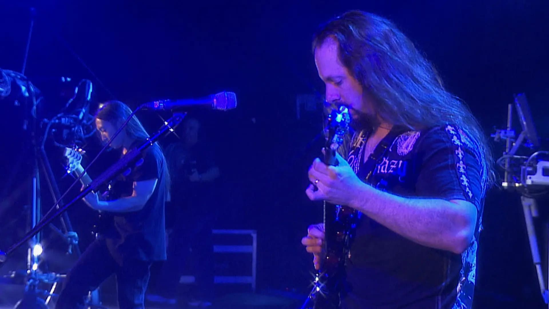 dream theater performing on stage with electric guitars under blue lighting. One is in the foreground focusing on his instrument, while the other is in the background. Microphones and equipment are visible.