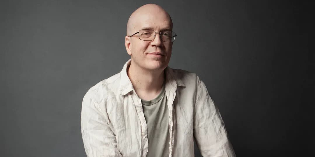Devin townasend in a light-colored shirt and gray t-shirt sits against a plain dark background.