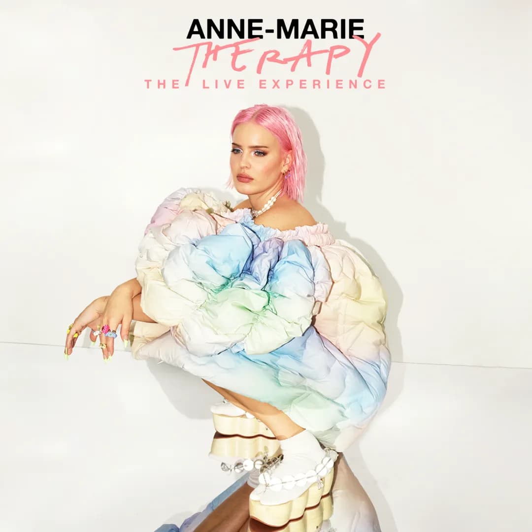Anne-Marie with pink hair in a colorful, puffy dress sits on a reflective floor. Text reads "Anne-Marie Therapy: The Live Experience.
