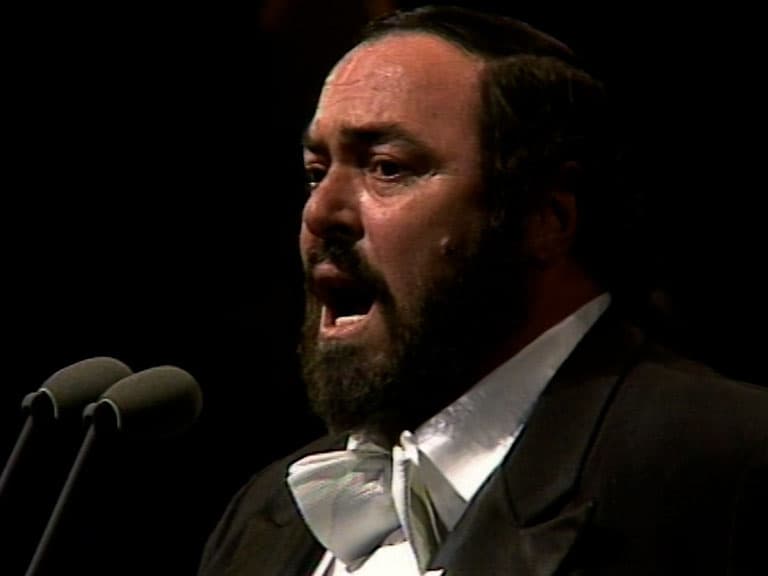 luciano pavarotti in formal attire is singing passionately into a microphone on stage.