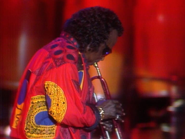 Miles Davis in a colorful shirt plays the trumpet on stage against a red backdrop.