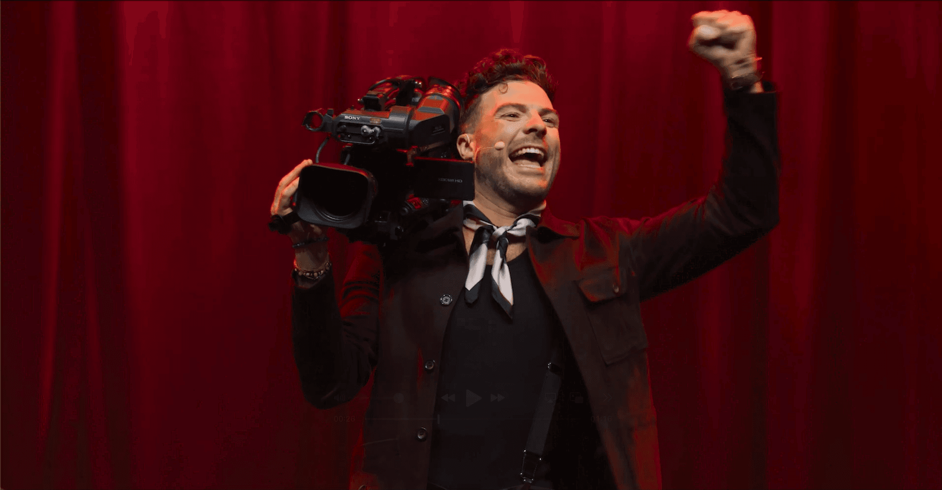 A man stands in front of red curtains, holding a video camera on his shoulder with one hand and raising his other fist in the air, appearing enthusiastic.