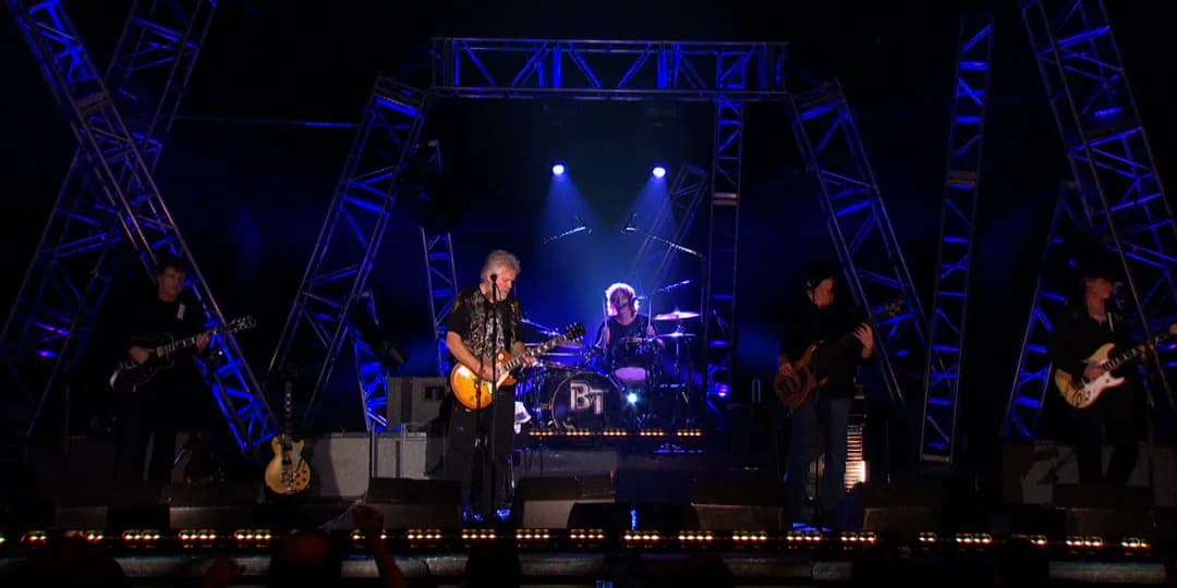 Bacjman & Turner performs on stage with blue lighting and metal truss structures; the drummer is centered, flanked by guitarists and a bassist.