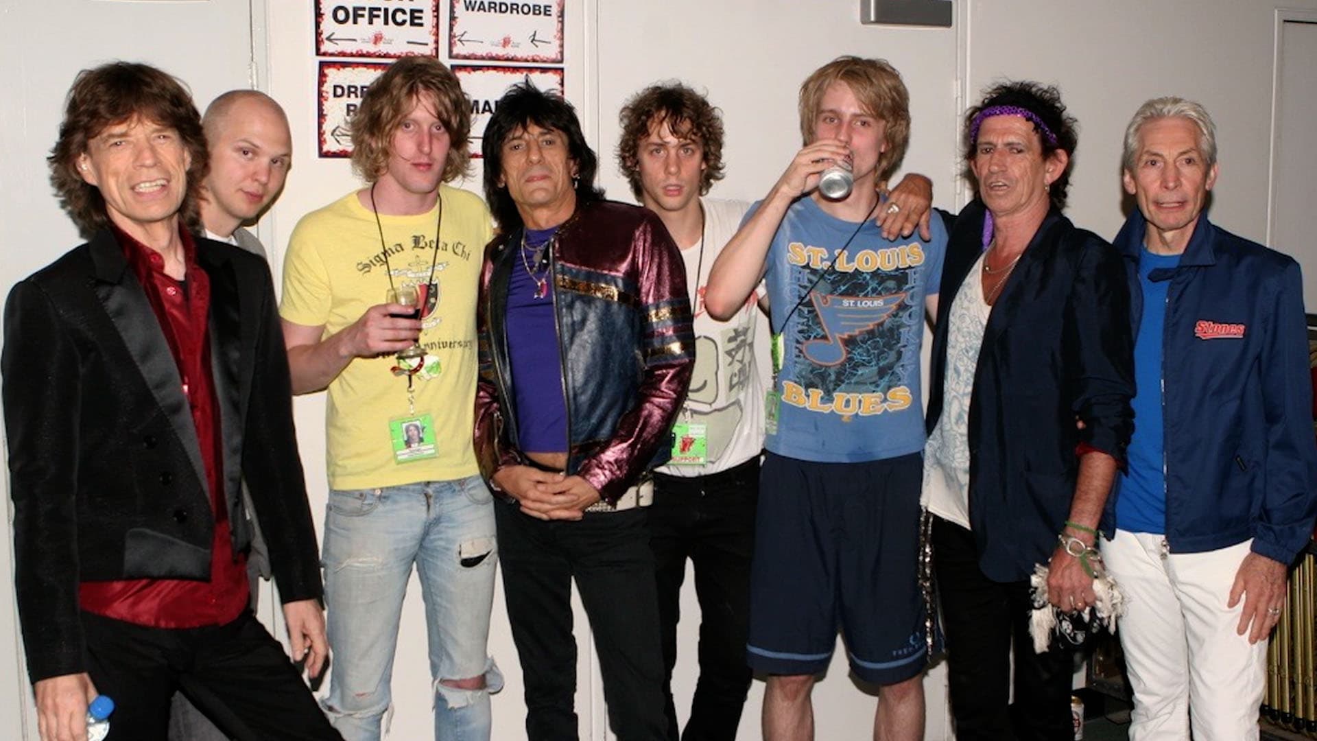Razorlight and The Rolling Stones standing together, some holding drinks. They are casually dressed, with varied hairstyles and band t-shirts. A few are smiling or looking at the camera.