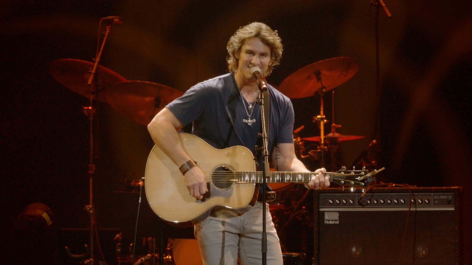 Dierks Bentley plays an acoustic guitar and sings into a microphone on stage, with drums and amplifiers visible in the background.