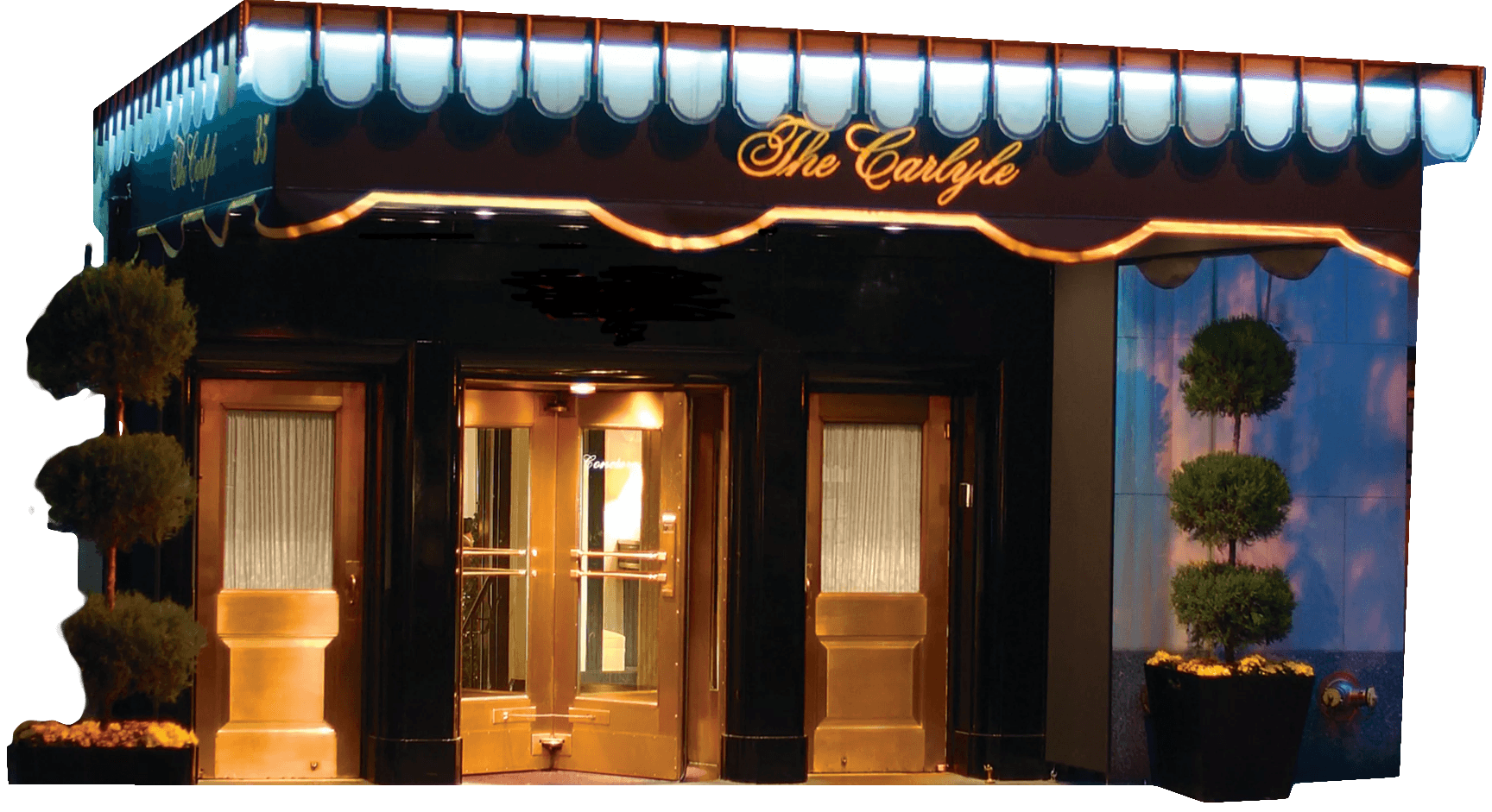 Entrance of The Carlyle hotel featuring a black and gold awning with illuminated signage, double glass doors, and manicured topiary plants on either side.