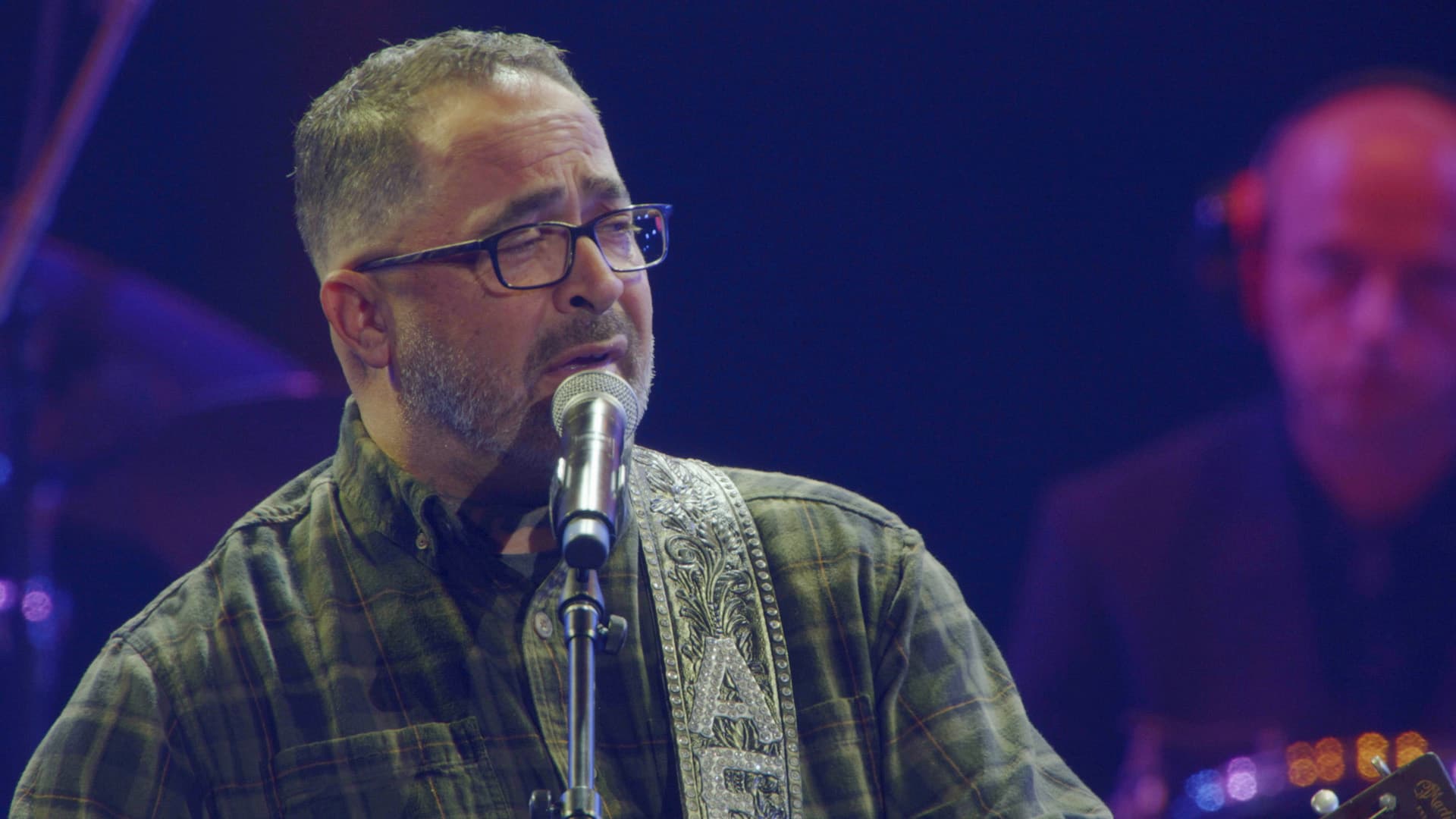 Aaron Lewis wearing glasses and a plaid shirt sings into a microphone on stage; a musician is visible in the blurred background.