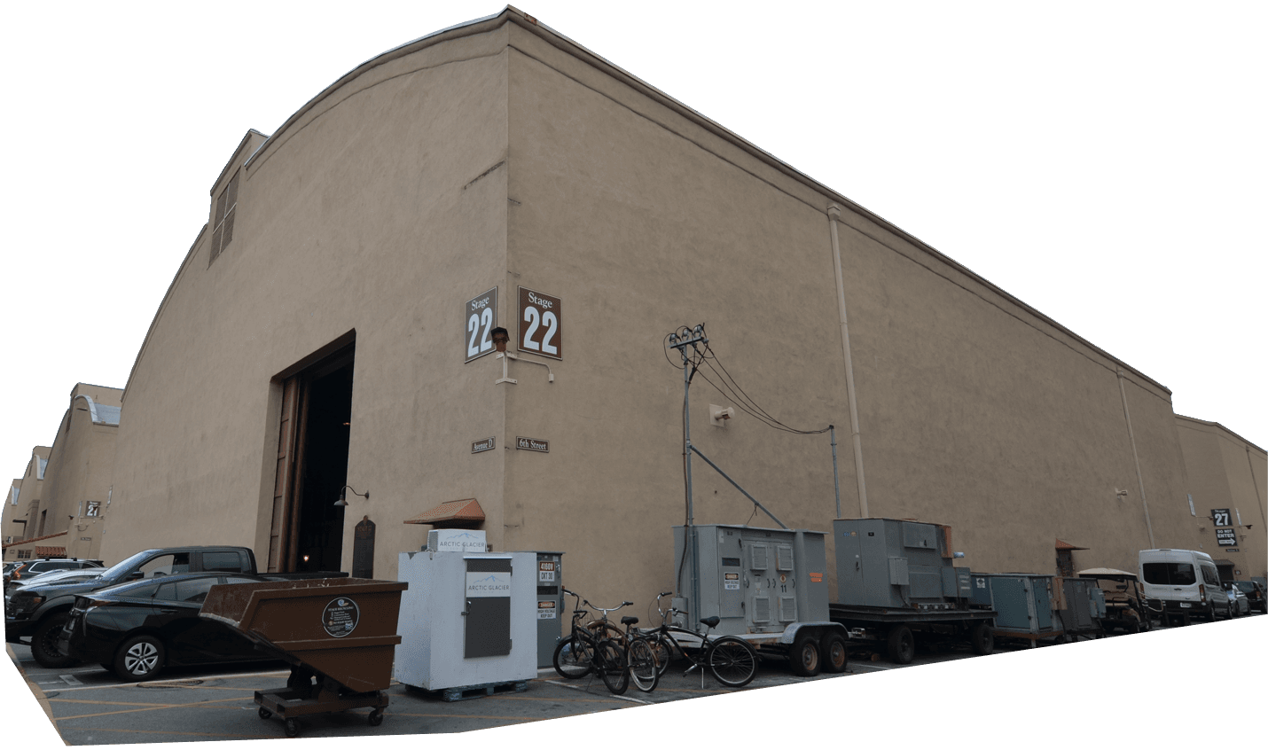 Large beige warehouse-style building labeled "Stage 22" with vehicles, dumpsters, bicycles, and equipment outside on an overcast day.