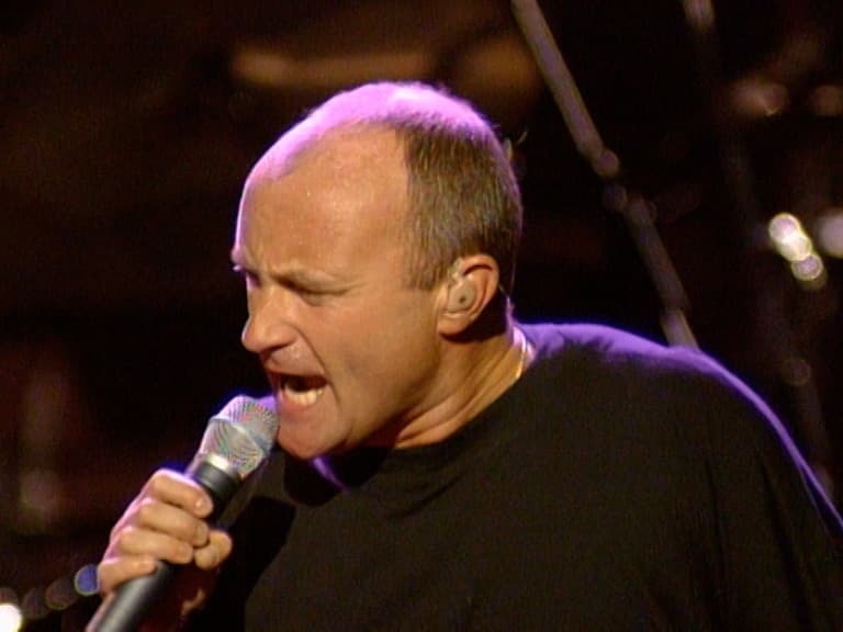 Phil Collins sings passionately into a microphone on stage, wearing a black shirt.