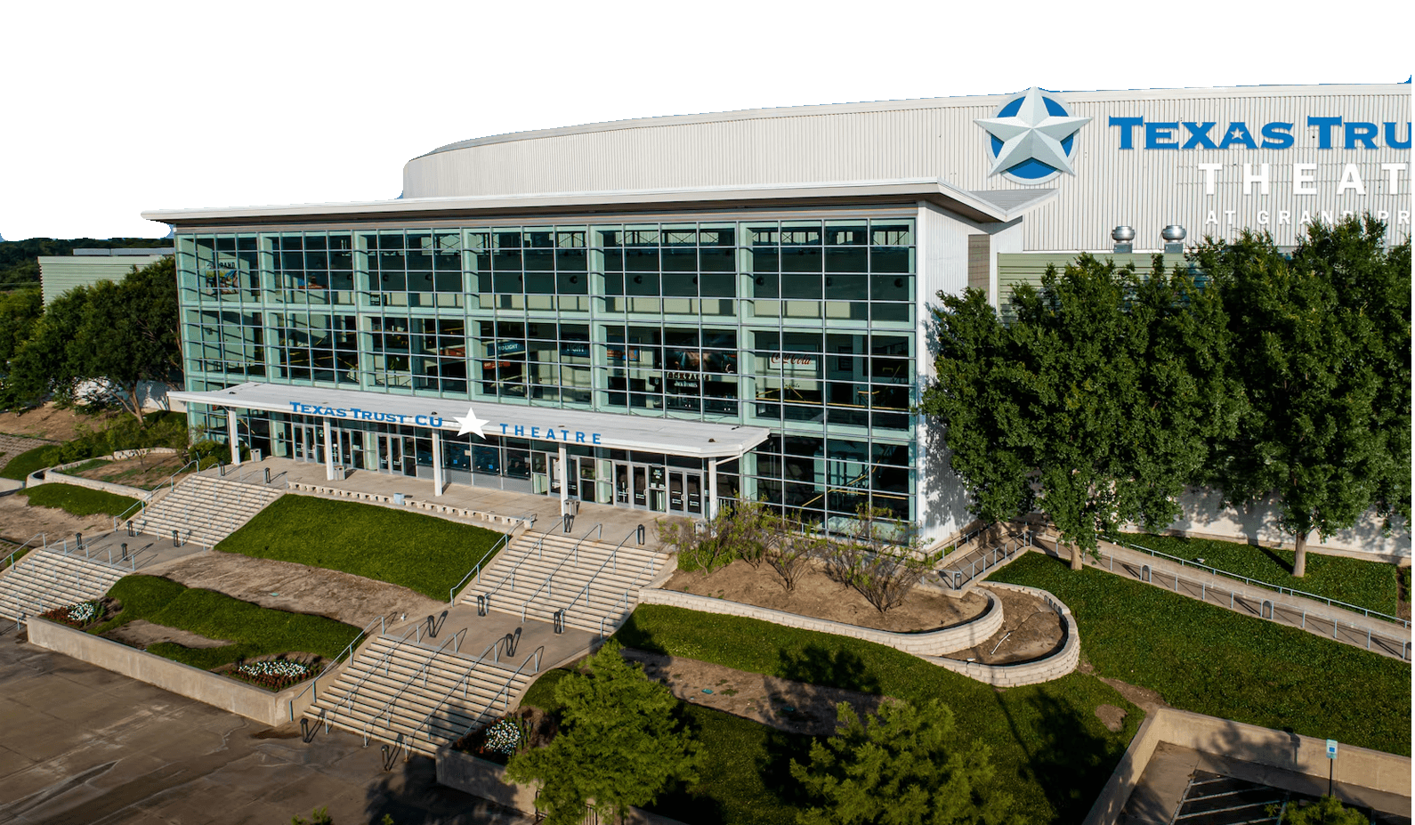 A modern glass-fronted building labeled “Texas Trust CU Theatre” with multiple entry doors, staircases, and landscaped greenery in front.