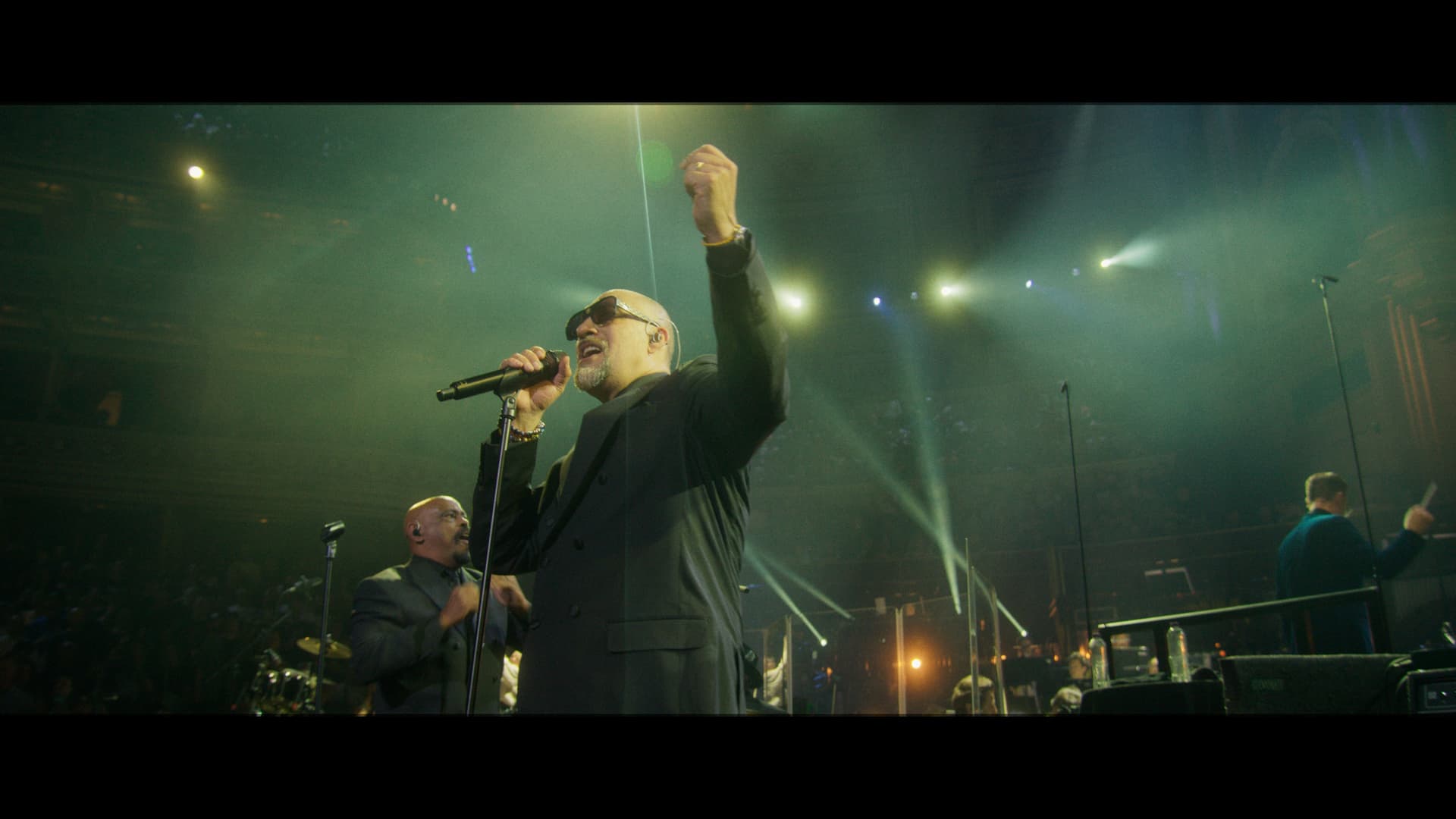 Cypress Hill singer wearing sunglasses performs on stage with a microphone, backed by a band and colorful stage lights in a large concert venue.