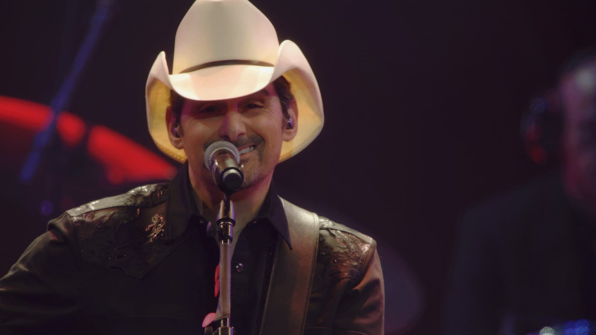 A man in a white cowboy hat and black shirt sings into a microphone on stage under dim lighting.