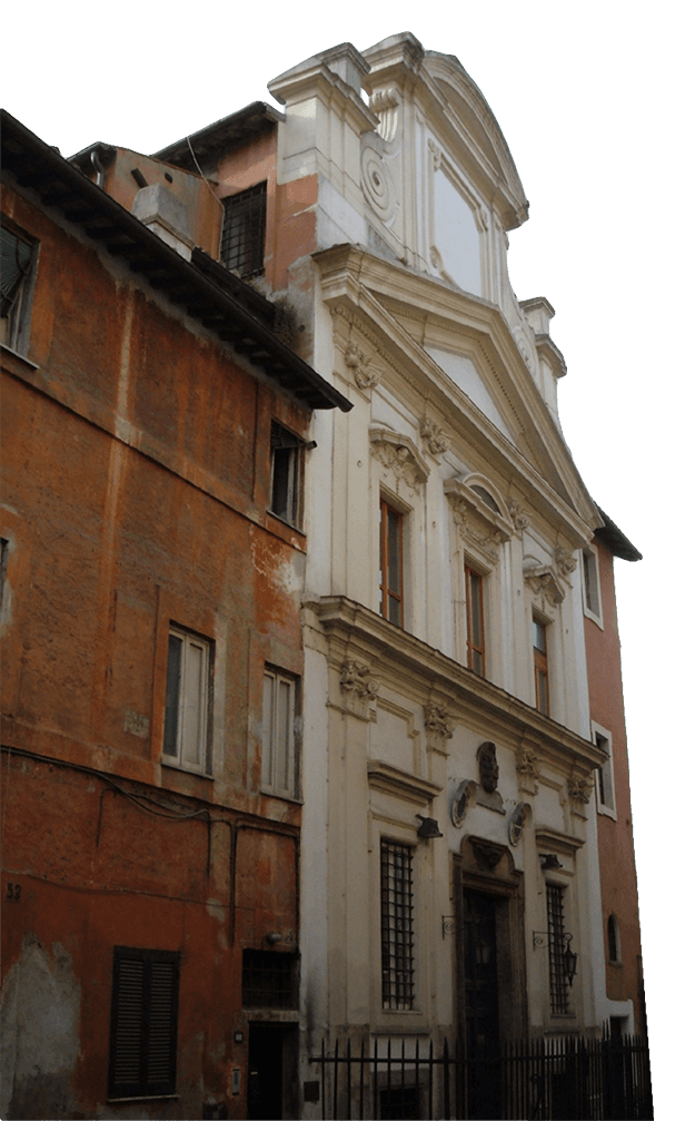 A white, baroque-style church facade stands between aged, reddish-brown residential buildings on a narrow urban street.