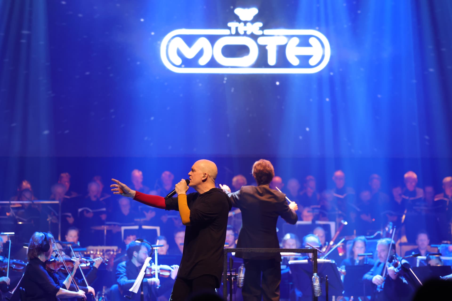 Devin Townsend sings on stage with an orchestra and conductor under blue lighting. The words "THE MOTH" are illuminated above. photographer: Wessel de Groot