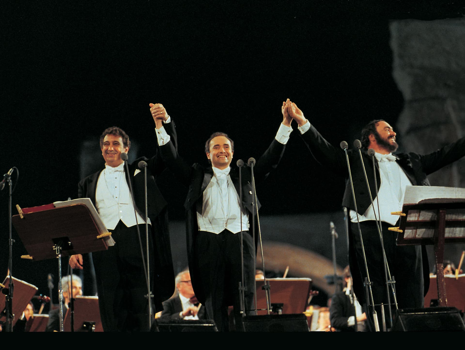 The Three Tenors in tuxedos stand on stage, holding hands and smiling, with music stands and an orchestra visible behind them.