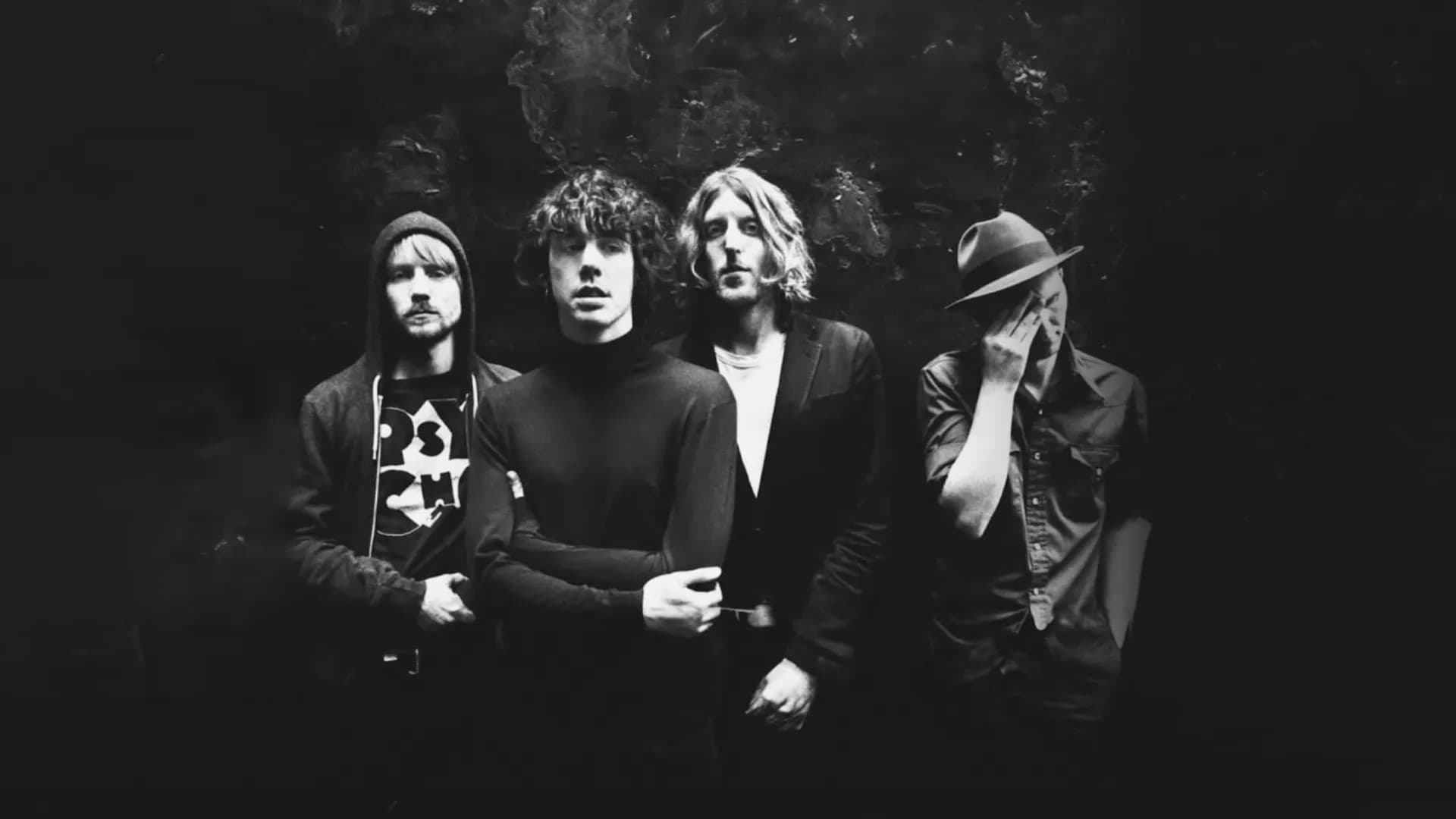 Razorlight stand against a dark background, one wearing a hat, another with curly hair, dressed in casual, dark clothing.