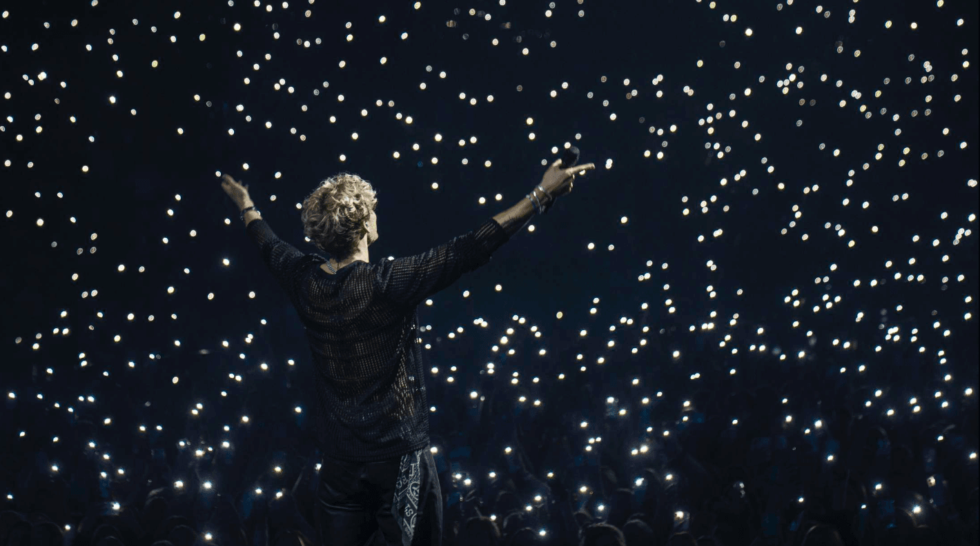 Christopher stands on stage with arms raised, facing an audience holding up lit phones in a dark concert venue.