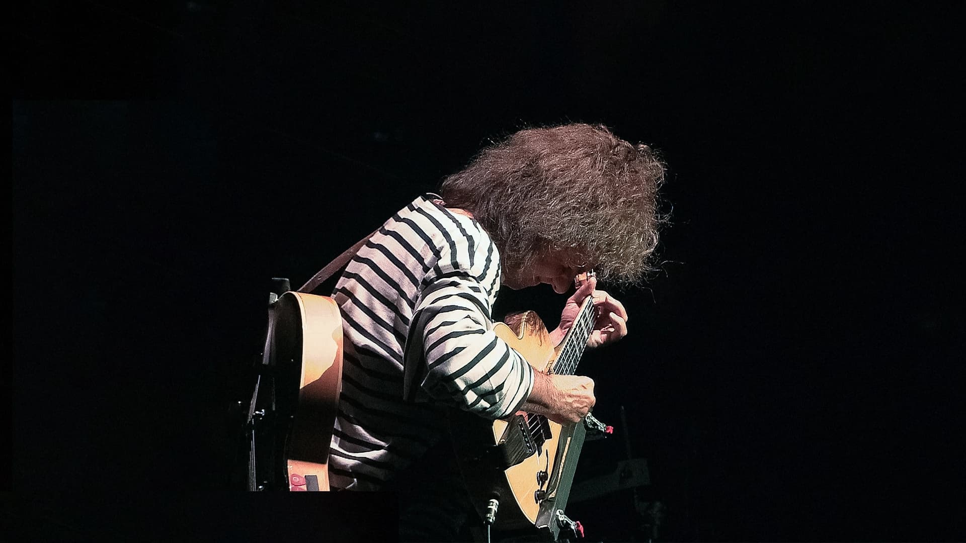 Pat Metheny playing an electric guitar on stage, wearing a striped shirt, with messy hair, in a dimly lit setting.