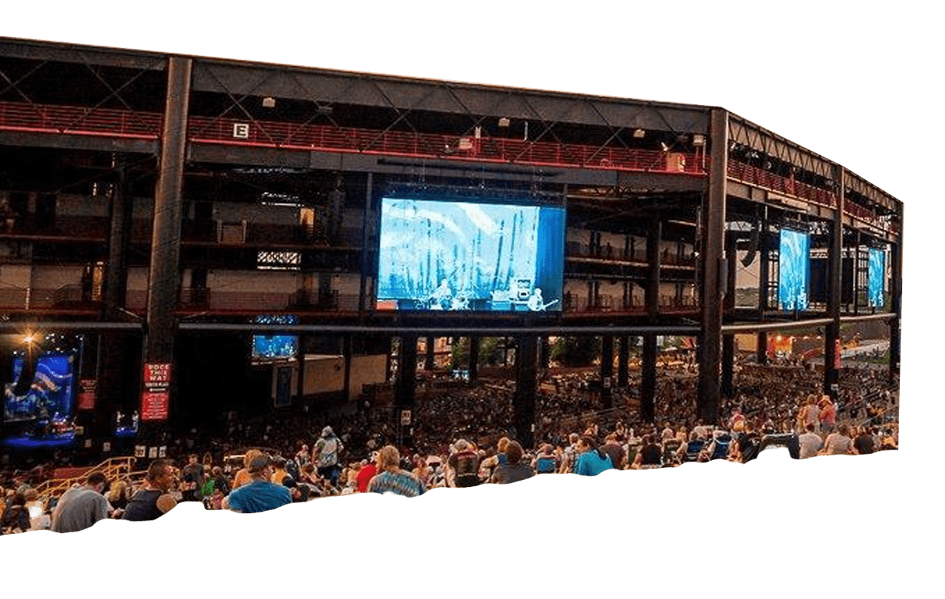 Large crowd seated in an open-air concert venue with multiple video screens displaying the stage, under a partially covered roof structure.