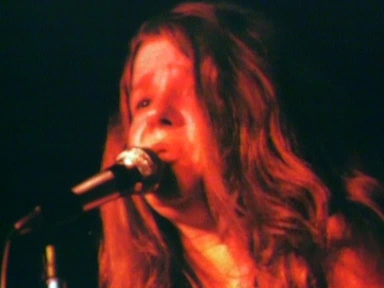 member of Big Brother & The Holding Company with long hair is singing into a microphone on stage under warm red lighting.