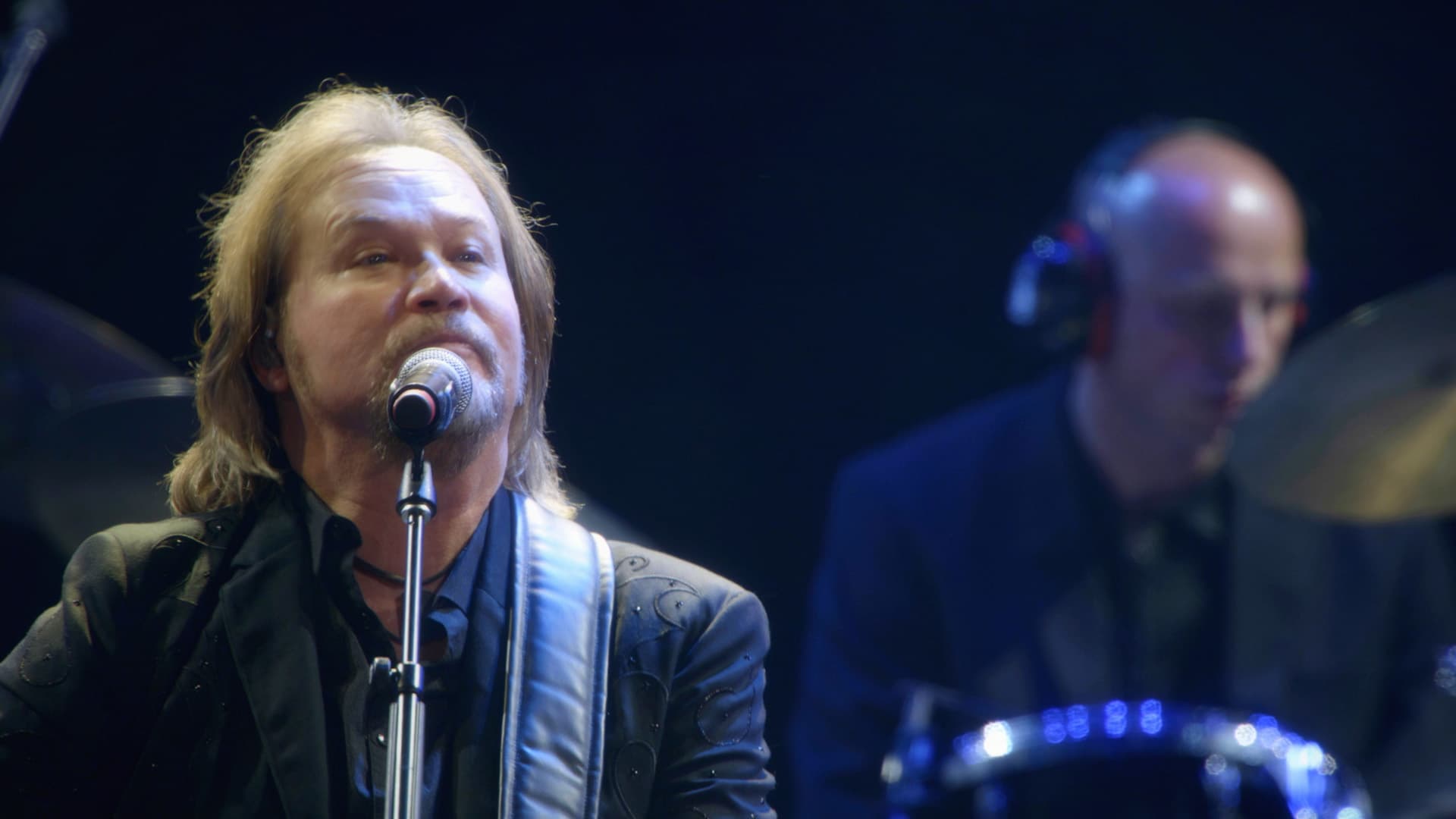 Travis Tritt sings into a microphone on stage, with a drummer playing in the background.